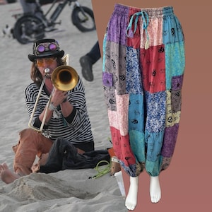 Pode incluir: Calças harem de patchwork multicoloridas com cintura com cordão. As calças são exibidas em um manequim branco. A imagem também mostra uma pessoa tocando trombone na praia. O patchwork inclui painéis azuis, vermelhos, roxos e rosa.