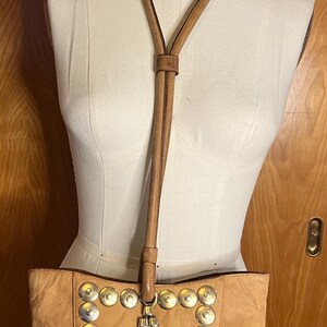 May include: A tan leather tote bag with a long strap and silver metal accents. The bag has a fringe detail and is decorated with round silver studs.