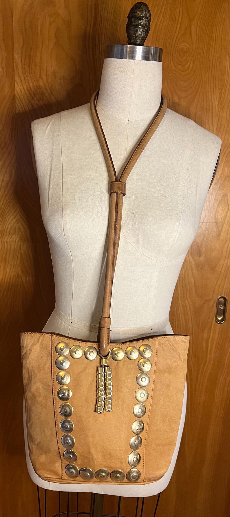 May include: A tan leather tote bag with a long strap and silver metal accents. The bag has a fringe detail and is decorated with round silver studs.