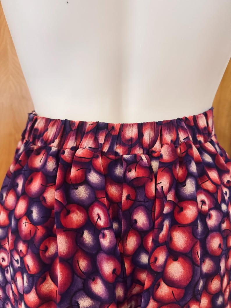 Peut inclure: Un pantalon taille haute avec un imprim&eacute; de cerises. Le tissu pr&eacute;sente un motif de cerises rouges et violettes avec des tiges vertes sur un fond violet fonc&eacute;. La taille est &eacute;lastique.