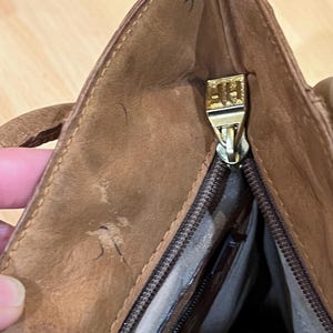 May include: A brown leather tote bag with a gold zipper and a gold pull tab. The zipper is open and the inside of the bag is visible.