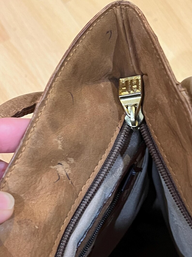 May include: A brown leather tote bag with a gold zipper and a gold pull tab. The zipper is open and the inside of the bag is visible.