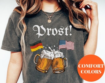 Könnte beinhalten: Dunkelgraues T-Shirt mit dem Wort "Prost!" in weißer Schrift. Das Shirt zeigt eine Grafik von zwei Bierkrügen, die anstoßen, mit deutschen und amerikanischen Flaggen. Die Worte "Comfort Colors" stehen in einem orangefarbenen Kreis.