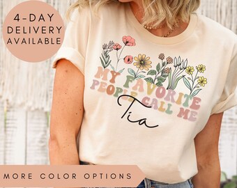 May include: A light beige t-shirt with a floral design and the text "My Favorite People Call Me Tia" in colorful letters.
