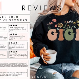 May include: Black sweatshirt with a floral design and the word "GIGI" in a retro font. The sweatshirt is being modeled by a person holding a white mug. The text "OVER 7000 HAPPY CUSTOMERS" and several five-star reviews are visible in the background.