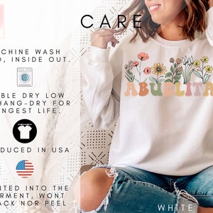 May include: White sweatshirt with a floral design and the text "Abuelita" in colorful letters. The sweatshirt is shown on a model wearing blue jeans. The text "CARE" is in black letters at the top of the image. The text "MACHINE WASH COLD, INSIDE OUT." is in black letters. The text "TUMBLE DRY LOW OR HANG-DRY FOR LONGEST LIFE." is in black letters. The text "PRODUCED IN USA" is in black letters. The text "PRINTED INTO THE GARMENT, WONT CRACK NOR PEEL" is in black letters. The text "WHITE" is in black letters.