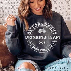 A woman wearing a gray sweatshirt with a white circular design that says "Oktoberfest Drinking Team" and "2026" printed on it, smiling and sitting on a couch.