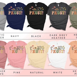 K&ouml;nnte beinhalten: Eine Sammlung von T-Shirts mit einem floralen Design und dem Text "PROST!" in einer Retro-Schriftart. Die Shirts sind in verschiedenen Farben erh&auml;ltlich, darunter Sport-Heather, Marine, Schwarz, Dunkelgrau-Heather, Heather Olive, Heather Mauve, Pink, Natur, Wei&szlig; und Heather Peach.