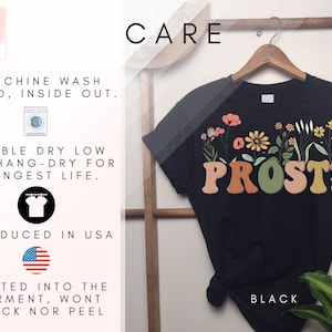 K&ouml;nnte beinhalten: Schwarzes T-Shirt mit floralem Design und dem Aufdruck "PROST!" auf der Vorderseite. Das Shirt ist in den USA hergestellt und maschinenwaschbar.