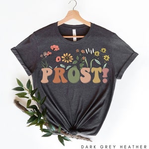 K&ouml;nnte beinhalten: Dunkelgraues T-Shirt aus Heather-Material mit einem floralen Design und dem Wort "PROST!" in bunten, Retro-Lettern.
