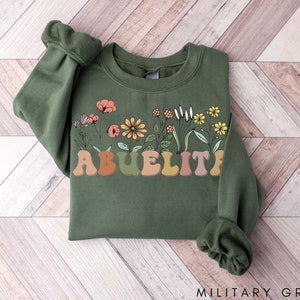 May include: A green sweatshirt with a floral design and the word "ABUELITA" in colorful, retro-style letters.