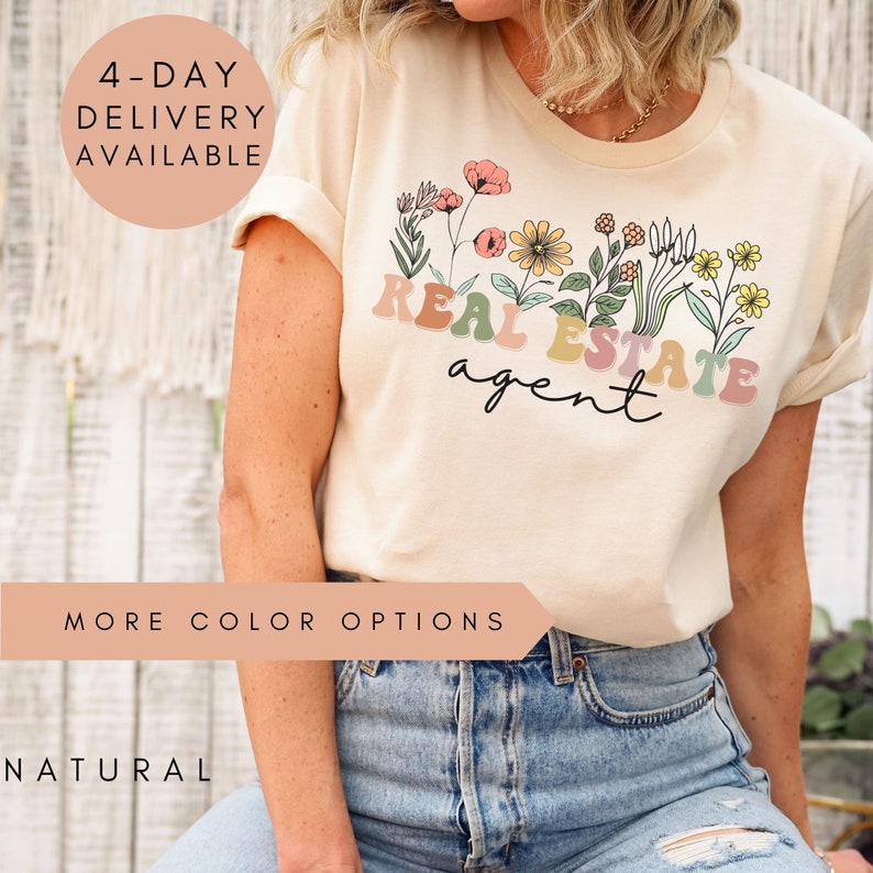 May include: A beige t-shirt featuring a floral design and the text "REAL ESTATE agent" in colourful letters. The shirt is being worn by a person with blue jeans.