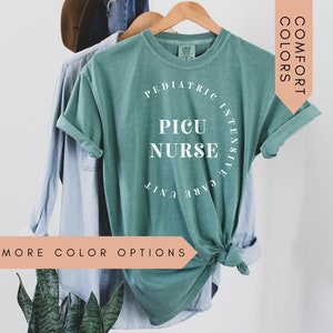 K&ouml;nnte beinhalten: Ein blaugr&uuml;nes T-Shirt mit dem Text "Pediatric Intensive Care Unit PICU Nurse" in Wei&szlig;. Das Shirt h&auml;ngt an einem Kleiderb&uuml;gel und hat einen Knoten vorne.