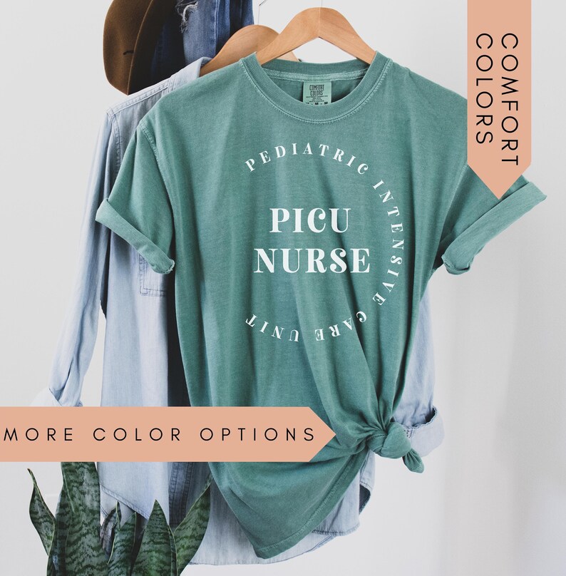 K&ouml;nnte beinhalten: Ein blaugr&uuml;nes T-Shirt mit dem Text "Pediatric Intensive Care Unit PICU Nurse" in Wei&szlig;. Das Shirt h&auml;ngt an einem Kleiderb&uuml;gel und hat einen Knoten vorne.