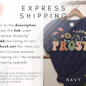 K&ouml;nnte beinhalten: Marineblaues T-Shirt mit floralem Design und dem Text "PROST!" in bunten Blockbuchstaben. Der Text "NAVY" ist in Wei&szlig; am unteren Rand des Shirts gedruckt.