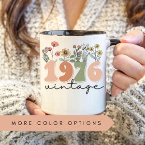 A person is holding a mug with the text "1976 Vintage" printed on it, surrounded by colorful floral designs.