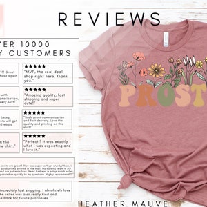 K&ouml;nnte beinhalten: Ein T-Shirt in Heidekraut-Mauve mit einem floralen Design und dem Wort "PROST" in bunten Buchstaben. Das Shirt ist auf einer wei&szlig;en Holzoberfl&auml;che abgebildet. Das Bild enth&auml;lt Kundenbewertungen, die die Qualit&auml;t, den Versand und die Personalisierung des Shops loben.