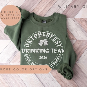 a green sweatshirt with a white circular design on it, which appears to be related to an Oktoberfest Drinking Team event. The text on the sweatshirt reads "OKTOBERFEST DRINKING TEAM" and "2026". The sweatshirt is laid out on a wooden surface, and there is a text overlay that says "EXPRESS SHIPPING AVAILABLE" and "MORE COLOR OPTIONS".