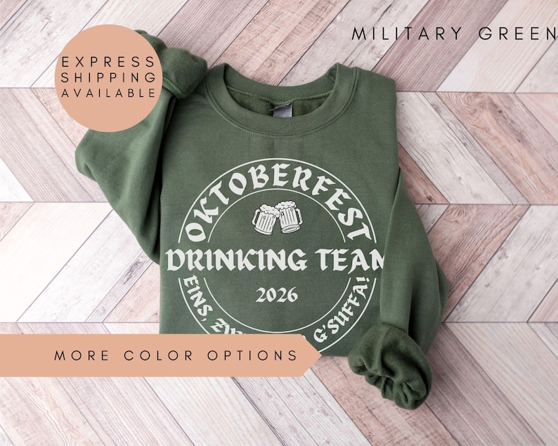 a green sweatshirt with a white circular design on it, which appears to be related to an Oktoberfest Drinking Team event. The text on the sweatshirt reads "OKTOBERFEST DRINKING TEAM" and "2026". The sweatshirt is laid out on a wooden surface, and there is a text overlay that says "EXPRESS SHIPPING AVAILABLE" and "MORE COLOR OPTIONS".