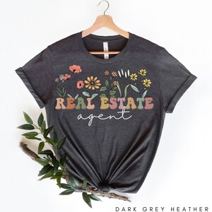 May include: Dark grey heather t-shirt with a floral design and the text "REAL ESTATE agent" in colourful letters.