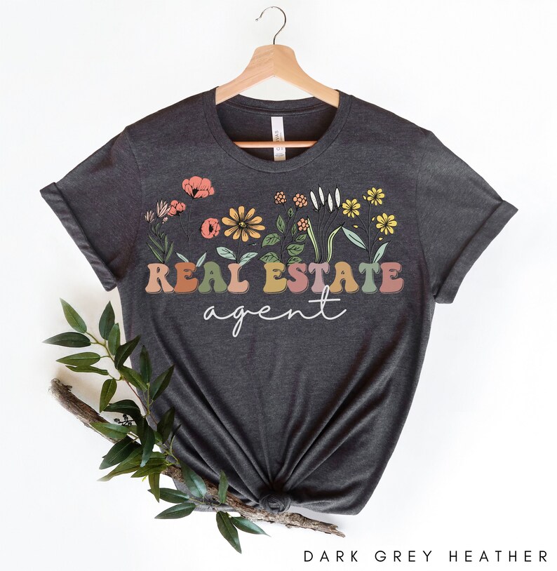 May include: Dark grey heather t-shirt with a floral design and the text "REAL ESTATE agent" in colourful letters.
