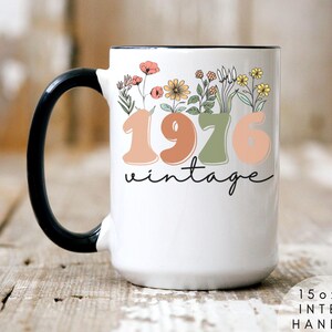 A white mug with a black handle, featuring a floral design and the text "1976 Vintage" printed on it.