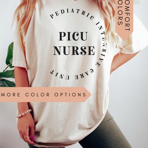 K&ouml;nnte beinhalten: Ein wei&szlig;es T-Shirt mit schwarzem Text, der "Pediatric Intensive Care Unit PICU Nurse" lautet. Der Text befindet sich in einem Kreis. Das Shirt wird von einer Person getragen. Der Text "Comfort Colors" befindet sich in einem pfirsichfarbenen Kasten in der oberen rechten Ecke des Bildes. Der Text "More Color Options" befindet sich in einem pfirsichfarbenen Kasten in der unteren linken Ecke des Bildes.