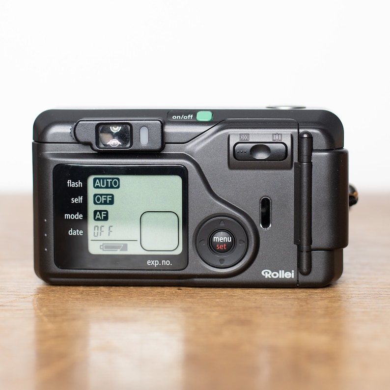 Może przedstawiać: Czarno-srebrny kompaktowy aparat fotograficzny z wyświetlaczem cyfrowym pokazującym ustawienia lampy błyskowej, samowyzwalacza, trybu i daty. Aparat ma przycisk menu i logo Rollei.