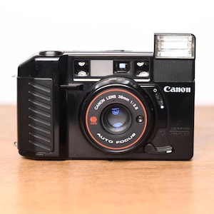 May include: A black Canon 35mm film camera featuring a 38mm 1:2.8 lens, an integrated flash unit on the top right, and an autofocus system. The front of the lens barrel displays the text Canon Lens 38mm 1:2.8, Auto Focus, and a red CAFS logo.