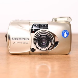 Könnte beinhalten: Eine champagnerfarbene Olympus Mju-III 120 Kompaktkamera mit einem 38-120mm Zoomobjektiv. Die Vorderseite zeigt den Markennamen, das Modell, einen ausklappbaren Blitz und einen blauen Aufkleber mit Wassertropfen-Symbol und Text ALL WEATHER.