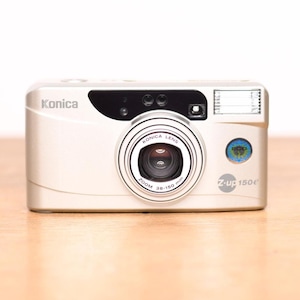 May include: A silver Konica Z-up 150e point-and-shoot film camera featuring a Konica Lens with 38-150 mm zoom capabilities. The front of the camera displays the Konica brand name, a black lens housing, a built-in flash, and a circular blue Eco sticker.