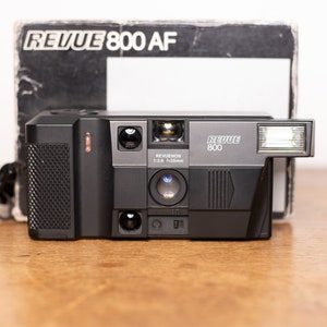 Puede incluir: Una cámara de película de 35 mm negra Revue 800 AF con flash incorporado. La cámara tiene una lente negra con el texto "Revueon 1:2.8 f=35mm" impreso en ella.