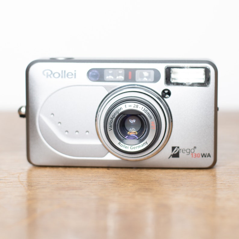 Może przedstawiać: Srebrny kompaktowy aparat fotograficzny Rollei Prego 130WA z obiektywem Vario-Apogon f=28-130mm. Aparat posiada wbudowaną lampę błyskową i srebrne wykończenie.