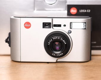 May include: A silver Leica C2 point-and-shoot film camera featuring a black Leica Vario-Elmar 35-70mm lens. The camera body displays the red Leica logo on the front, a viewfinder, a flash unit, and is positioned in front of its original box.