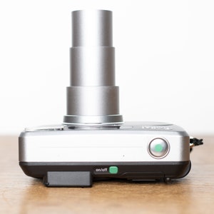 Może przedstawiać: Srebrny aparat cyfrowy z chowanym obiektywem i zielonym przyciskiem włączania/wyłączania. Aparat ma czarny pasek.