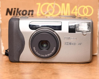 May include: A silver Nikon Zoom 400 AF camera with a black lens and flash. The camera has the Nikon logo and Zoom 400 AF printed on the front. The camera is on a wooden surface with a cardboard box in the background.