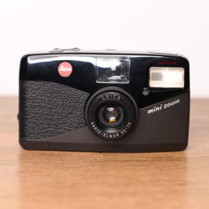 Könnte beinhalten: Eine schwarze Leica Mini Zoom 35-70 Kompaktkamera. Die Vorderseite verfügt über einen strukturierten Griff, ein rotes Leica-Logo, ein zentrales Objektiv mit der Aufschrift Vario-Elmar 35-70, ein Sucherfenster und einen rechteckigen Blitz oben rechts.