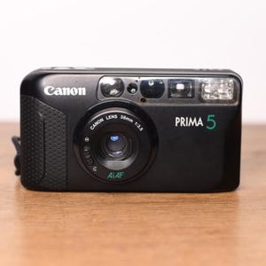 以下が含まれることがあります： 38mm f/3.5レンズとAiAF技術を搭載した黒いCanon Prima 5フィルムコンパクトカメラ。カメラの前面にはCanonのロゴ、緑色の文字でPrima 5というモデル名、内蔵フラッシュ、およびテクスチャ加工されたグリップエリアが表示されています。
