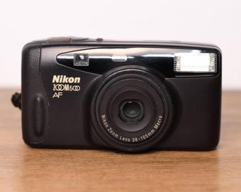 May include: A black Nikon Zoom 500 AF point-and-shoot camera featuring a Nikon Zoom Lens 38-105mm Macro. The front of the camera displays the brand name, model, and lens specifications in white text, along with a built-in flash and viewfinder.
