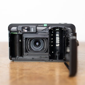 Może przedstawiać: Czarno-srebrny aparat fotograficzny 35 mm z otwartym komorą filmu, ukazujący kasetę filmu i obiektyw. Aparat ma zielony przycisk "on/off" i etykietę "film tip".