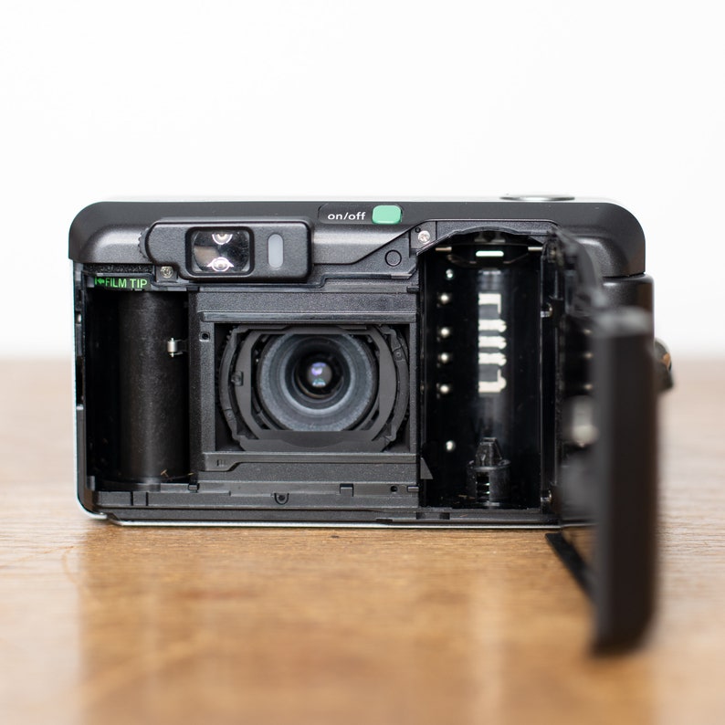 Może przedstawiać: Czarno-srebrny aparat fotograficzny 35 mm z otwartym komorą filmu, ukazujący kasetę filmu i obiektyw. Aparat ma zielony przycisk "on/off" i etykietę "film tip".