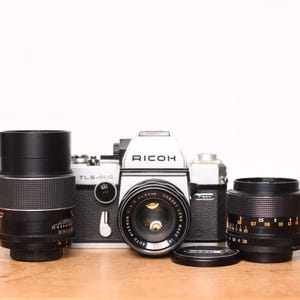 Puede incluir: Una cámara Ricoh vintage con dos lentes negras y una tapa de lente. El cuerpo de la cámara es plateado y negro, con el logotipo de Ricoh. Las lentes tienen marcas de medición.