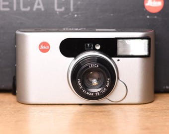 May include: A silver Leica C1 point-and-shoot camera featuring a Vario-Elmar 38-105 ASPH lens, a built-in flash, and the red Leica logo on the front. The camera is positioned in front of its original black box with the text LEICA C1 visible.