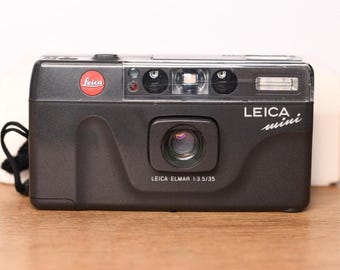 May include: A black Leica Mini point-and-shoot film camera featuring a red Leica logo, a central lens, and a built-in flash. The front of the camera displays the text Leica mini and Leica Elmar 1:3.5/35. A black wrist strap is attached to the side.