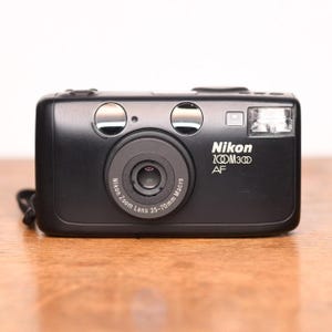 May include: Black Nikon Zoom 300 AF camera with a 35-70mm macro lens. The camera has a built-in flash and two silver buttons on top. The Nikon logo and model details are printed in white on the front.