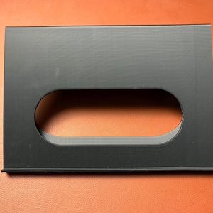 May include: A rectangular black plastic object with a large, oval-shaped cutout in the center. The object has a smooth, matte finish and appears to be a component or part of a larger item.