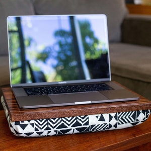 May include: A laptop computer sits on a wooden tray atop a black and white patterned cushion. The laptop is open and the screen shows a blurred image of trees and sky.