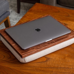May include: A laptop computer sits on a wooden tray with a white cushion. The tray is resting on a wooden table.