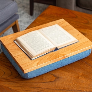 May include: A wooden tray with a blue cushion sits on a wooden table. A book is placed on the tray. The tray is perfect for reading in bed or on the couch.