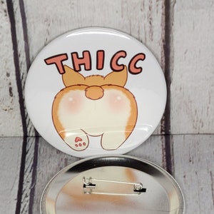 May include: A white button with a cartoon image of a corgi's rear end with the word "THICC" printed above it. The button has a silver back with a safety pin.
